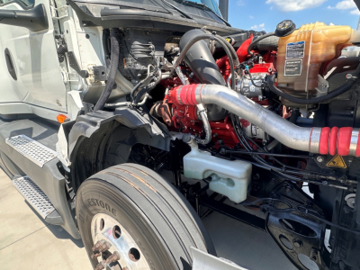 Truck engine bay of a 2021 International LT625 with visible coolant reservoir, intake piping, red engine block, and front tire, showing components and hose connections.