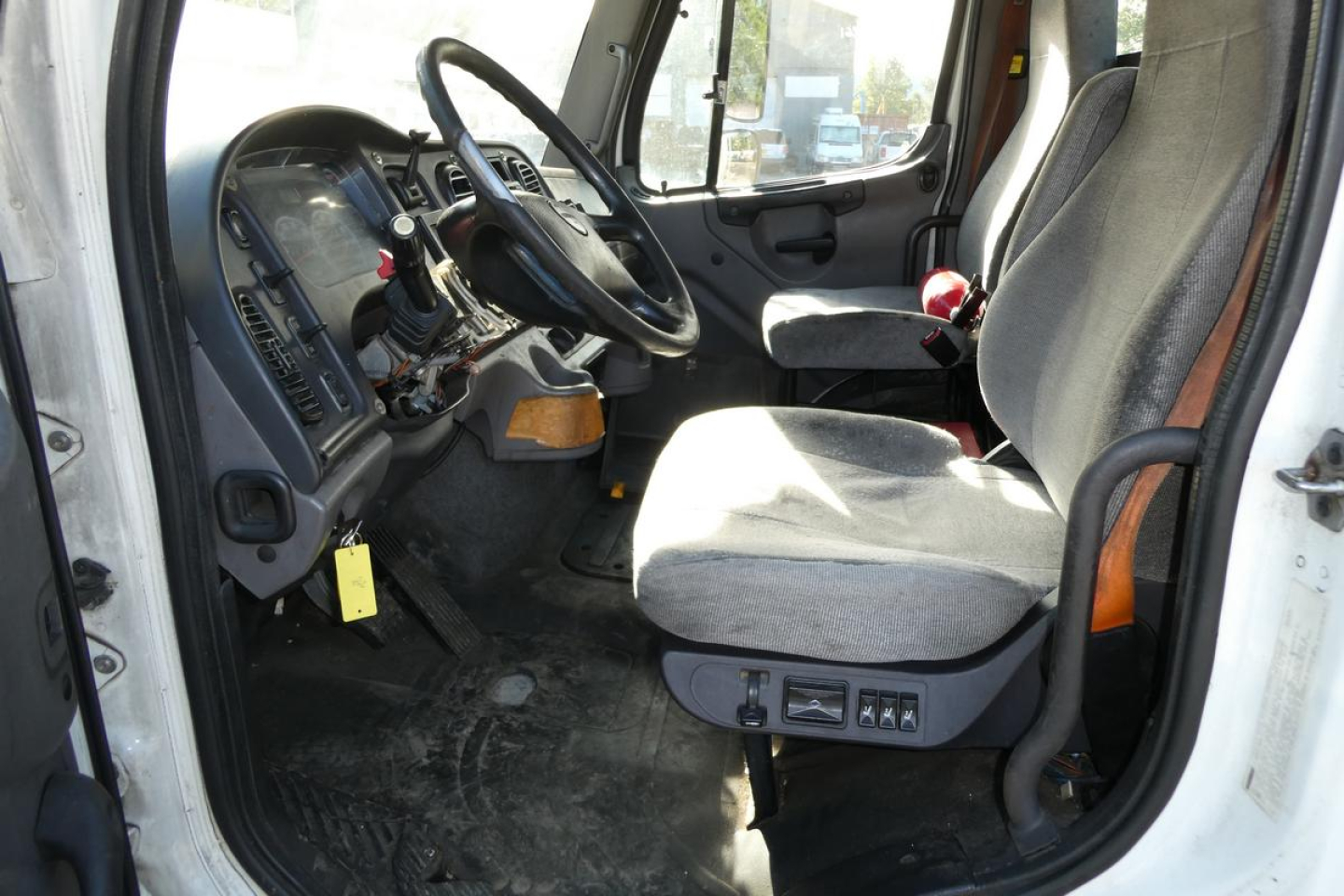Truck cab interior of a 2015 Freightliner M2 112 daycab showing steering wheel, dashboard, driver seat with controls, and pedals.