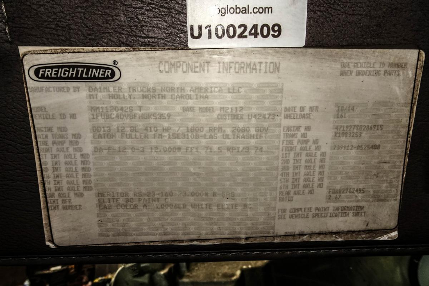 Freightliner component information plate with text blocks including model and VIN reference details. Visible words: 'FREIGHTLINER', 'COMPONENT INFORMATION', 'M2', multiple lines of small gray text, and a VIN-like sequence on a label nearby.