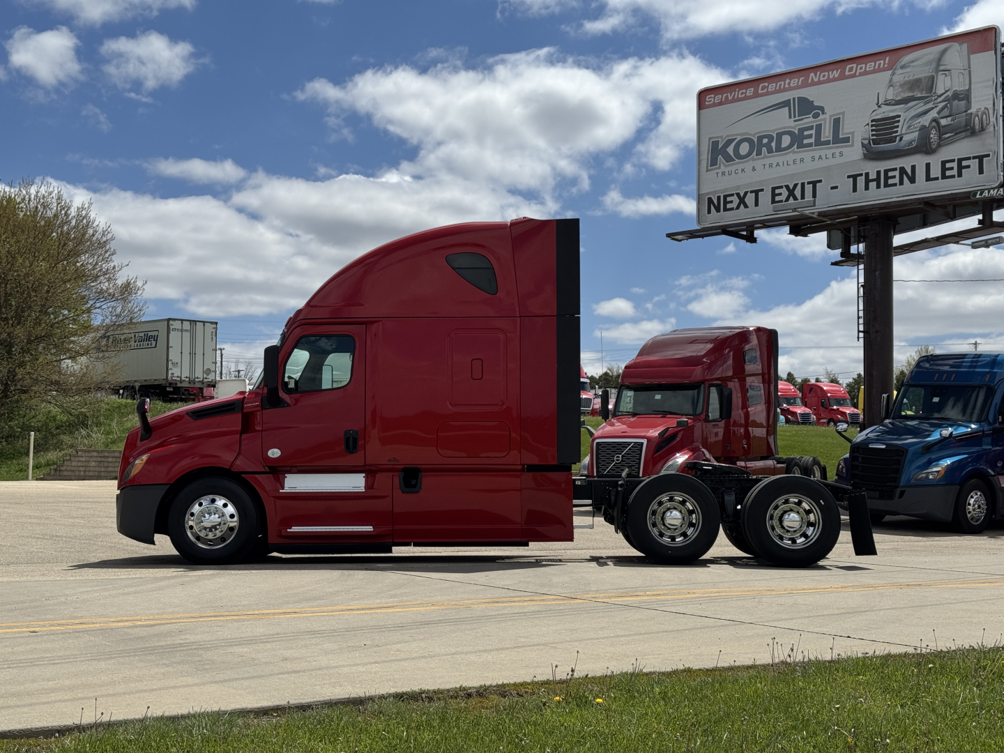 Freightliner Cascadia 126 truck tractor in red, side view with sleeper cab, tandem rear axles, dealership lot in background