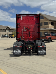 Freightliner Cascadia rear view showing red cab, wiring harness, and dual rear mud flaps with dealership stickers; visible rear bumper and chassis.