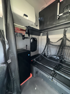 Freightliner Cascadia truck sleeper area with bunk bed and storage netting; compact fridge visible; interior panels and seat harnesses.