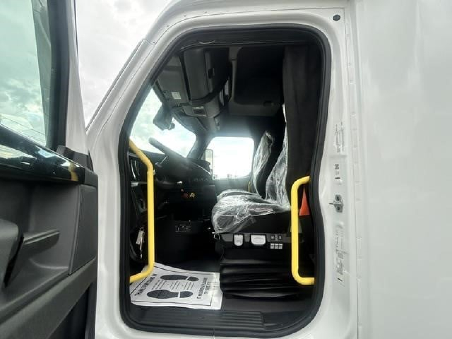 Truck cab interior of a 2024 Freightliner Cascadia 126, open driver door showing seat, steering wheel, and dashboard with yellow handrails.