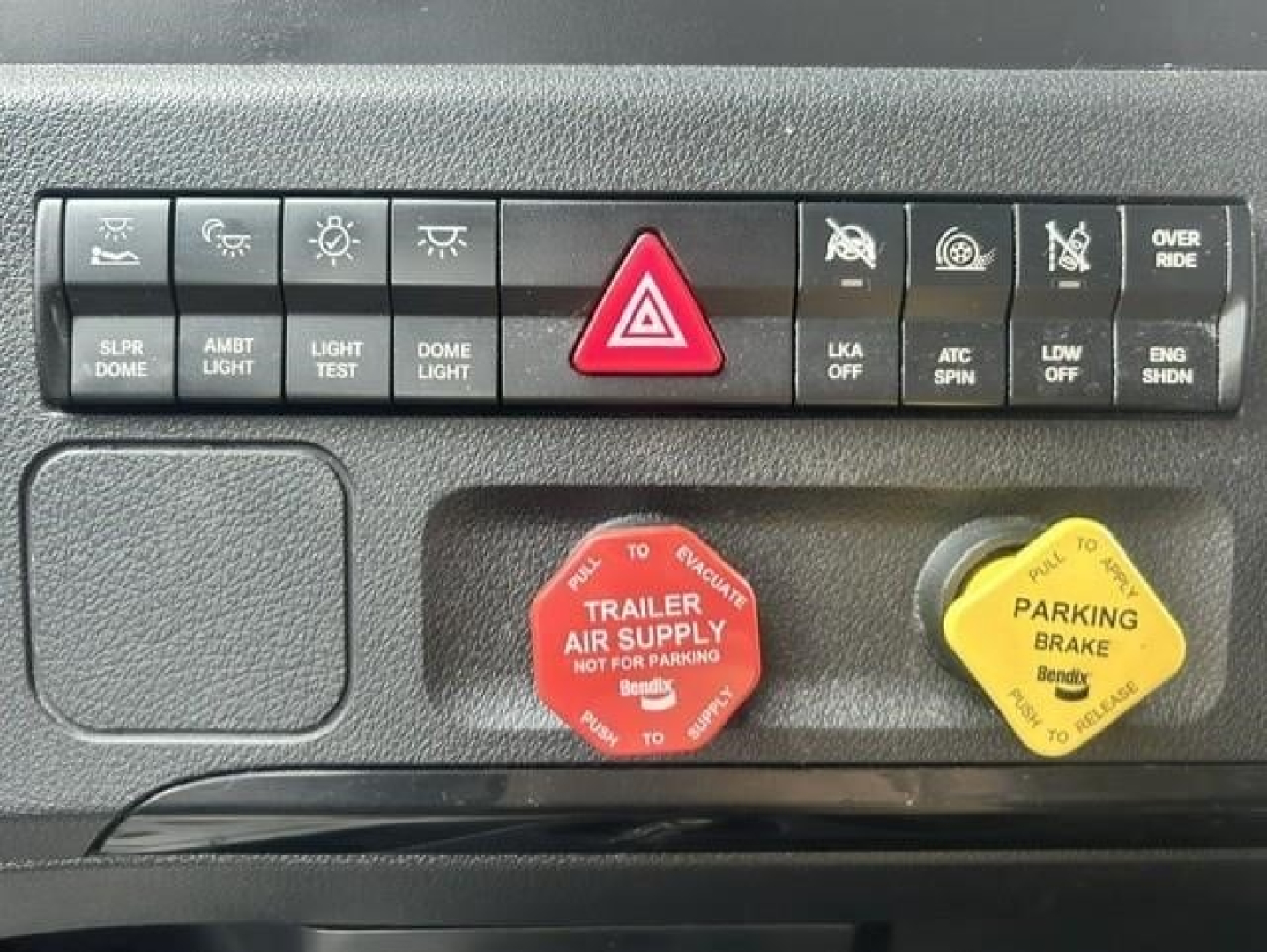 Dashboard switches and reservoir caps in a Freightliner Cascadia cab: red 'Trailer Air Supply' knob, yellow 'Parking Brake' knob, hazard button, various control icons.