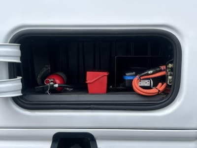 Truck storage compartment showing battery box, red plastic battery cover, orange jumper cables, and a red top tool box inside the side compartment.