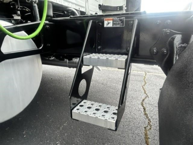 Truck undercarriage showing aluminum diamond-plate step/ladder attached to Freightliner Cascadia frame, fuel tank on left, green hose nearby, black chassis, visible rivets and brackets.