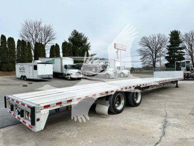 Trailer in showroom lot showing long flatbed with dual rear axles and drop deck, license plate not visible, no text readable other than small dealership signage in background.