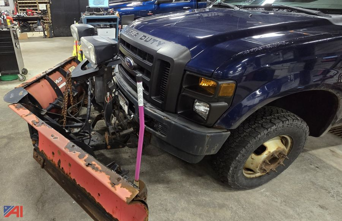 Ford F-350 Super Duty pickup with a front plow attachment mounted, in a workshop. Visible bumper, grille, blue cab, single front wheel, and snow plow blade showing wear.
