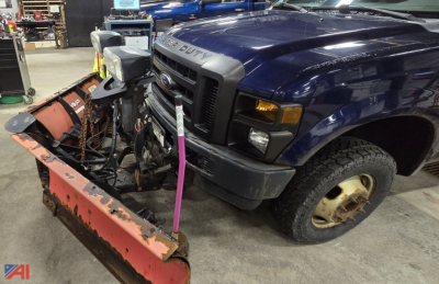 Ford F-350 Super Duty pickup with a front plow attachment mounted, in a workshop. Visible bumper, grille, blue cab, single front wheel, and snow plow blade showing wear.