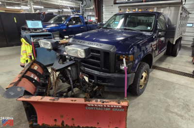Ford F-350 Super Duty dump truck in a shop with a plow attachment mounted at the front, blue cab, gray dump bed, indoor setting.