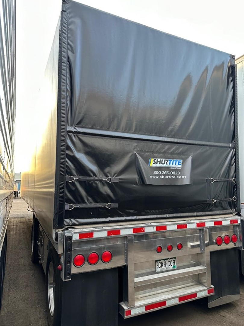 Trailer back with Conestoga sliding tarp system, black tarpaulin and tail gate, rear lights, license plate visible, reflective safety stripes.