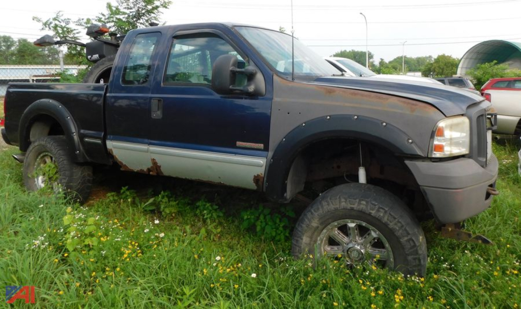 Ford F-350 Super Duty extended cab with plow, blue with beige underbody, raised suspension, large off-road tires, cargo bed, visible rust and dirt.