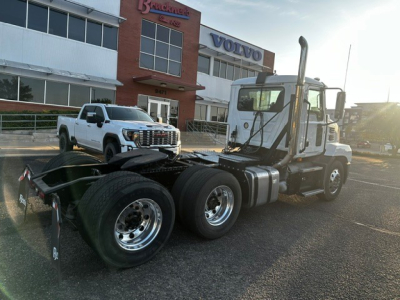 Truck tractor side/front view showing Mack Anthem cab with dual rear axles and fifth wheel, aluminum fuel tanks, exhaust stack, and showroom building in background.