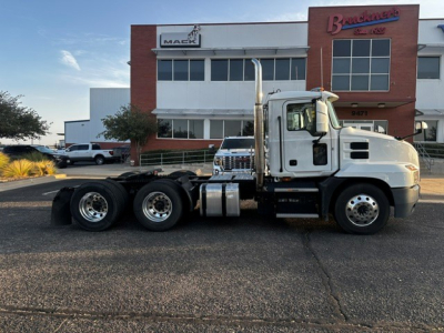 Side view of a white Mack Anthem semi-truck tractor chassis with three rear axles parked in front of a dealership building.