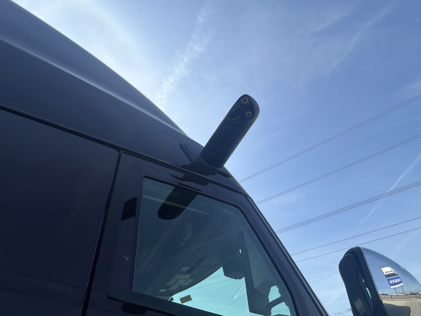 Mack PR64T76H truck cab corner with aerodynamic roof fairing and antenna visible, windshield and side window, blue sky.
