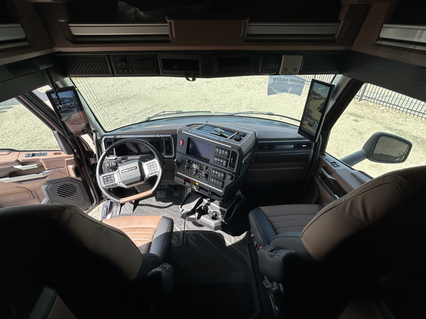 Truck cab interior showing Mack PR64T76H dashboard and steering wheel, with driver and passenger seats, large windshield, side mirrors, and electronic controls.