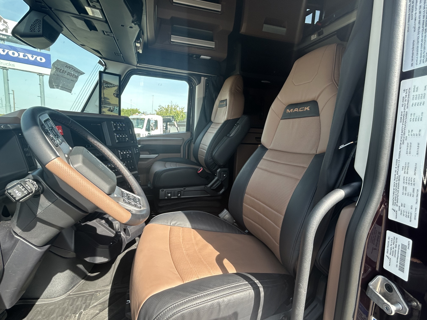 Truck cabin interior of a 2027 Mack PR64T76H showing tan and black driver and passenger seats, steering wheel, dashboard, and door frame.