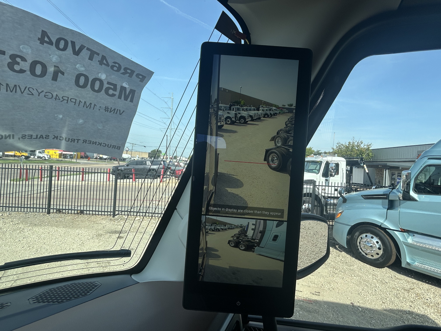 Truck cab interior with a vertical LCD monitor mounted at the windshield showing a reflection of a truck yard. Exterior scene includes other trucks and a banner reading 'PRE-TA.. M500 1...'
