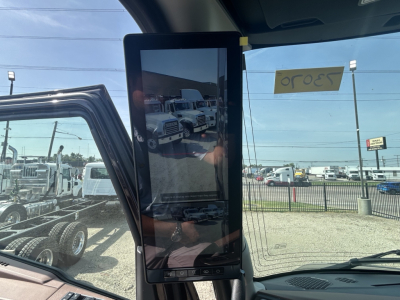 Large tablet-style display mounted inside Mack PR64T76H truck cab showing reflection of a used truck lot; no legible serials or fluid labels visible.