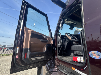 Truck cab interior view with open driver door showing dashboard and steering wheel of a Mack PR64T76H conventional sleeper in a maroon color.