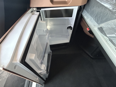 Small fridge/freezer in cabin with open door showing empty shelves and door bins; compact kitchenette area inside a truck, with light interior.
