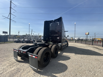 Mack PR64T76H conventional sleeper tractor rear view showing third-axle chassis and rear fenders with MAEK mud flaps; black exterior, high sleeper cab, parked on gravel lot.