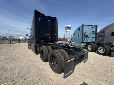 Mack PR64T76H truck chassis in a dealership lot showing rear view with dual rear axles and mud flaps, black sleeper cab in background.