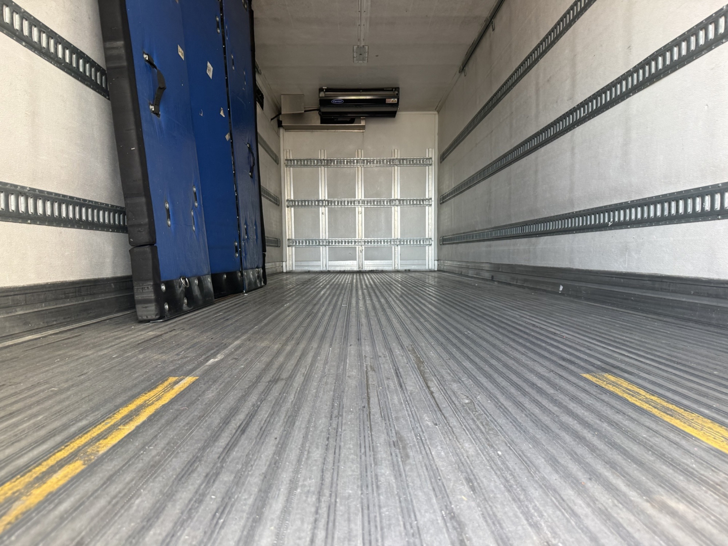 Interior of a refrigerated box truck trailer showing ribbed floor, sidewall tracks, and a blue mechanical unit near the left wall.