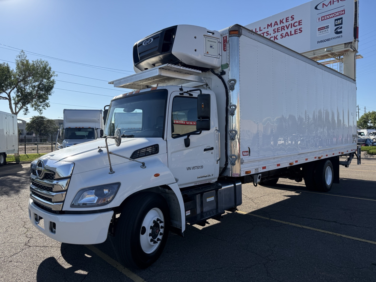 Hino 338 refrigerated box truck with a mounted freezer unit on top, white cab and cargo box, in a dealership lot.