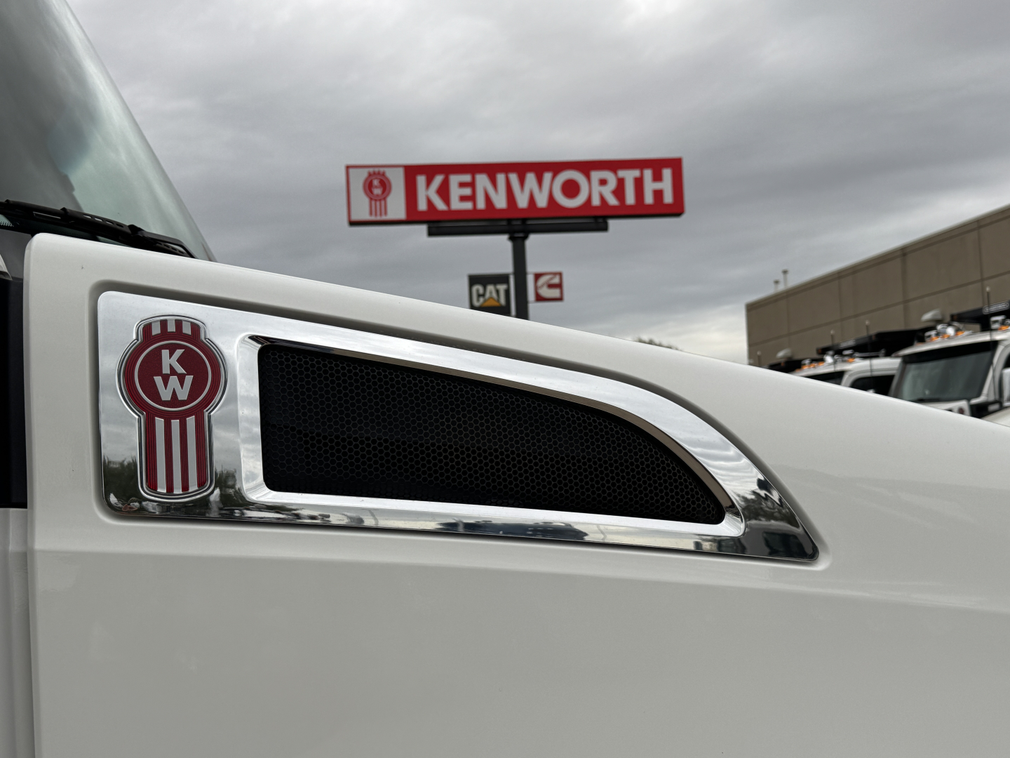 Kenworth emblem on white truck cab with black vent grille and Kenworth dealership sign in background.