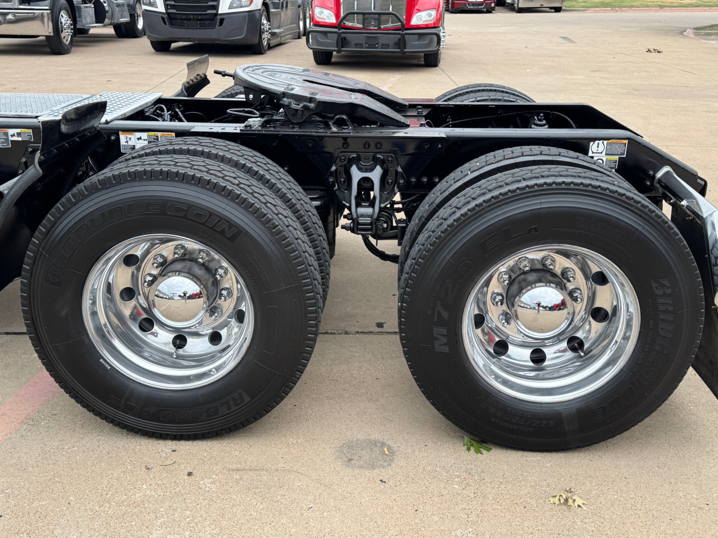 Kenworth T680 wheel area showing dual rear axles with chrome wheels and bold tires, chassis frame, and suspension components visible.