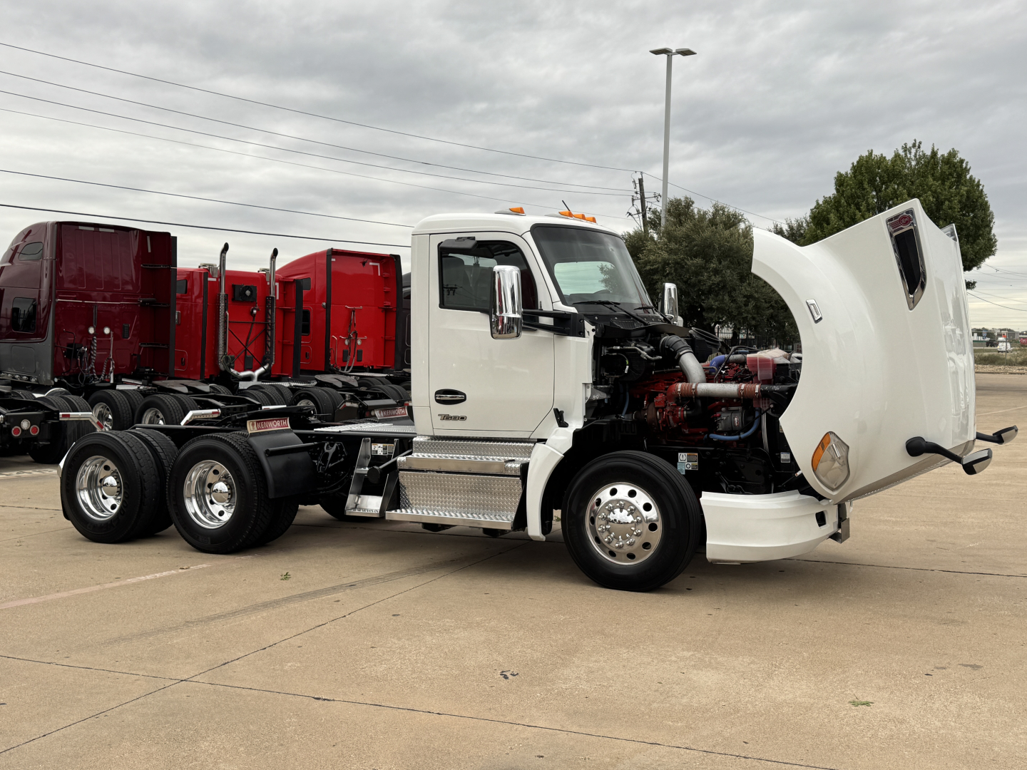 Kenworth T680 cab over with hood open showing engine and front wheel; white cab, multiple red trailers in background.