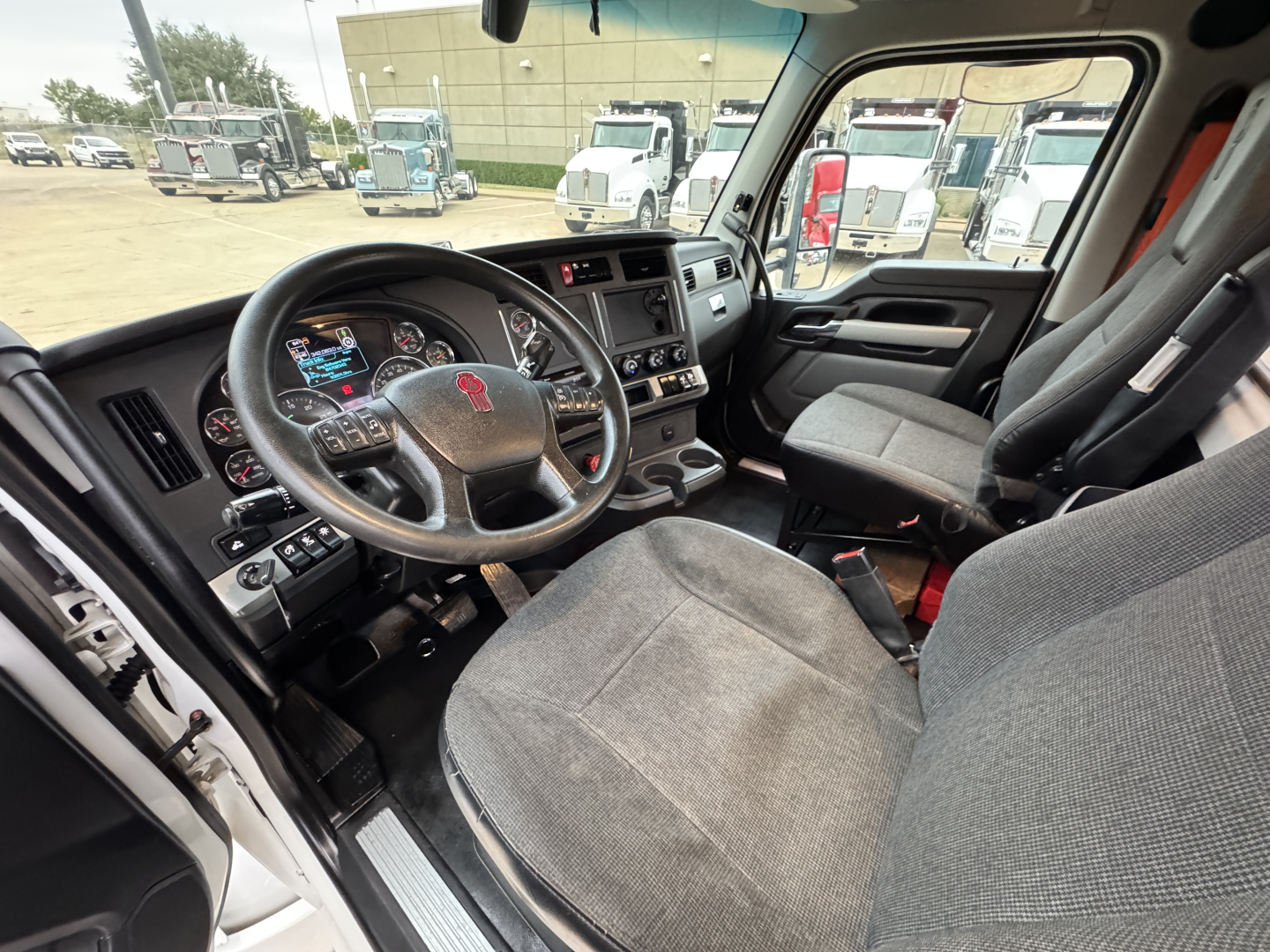 Kenworth T680 truck interior showing steering wheel with logo, dashboard, and driver seat; front passenger seat and center console visible.