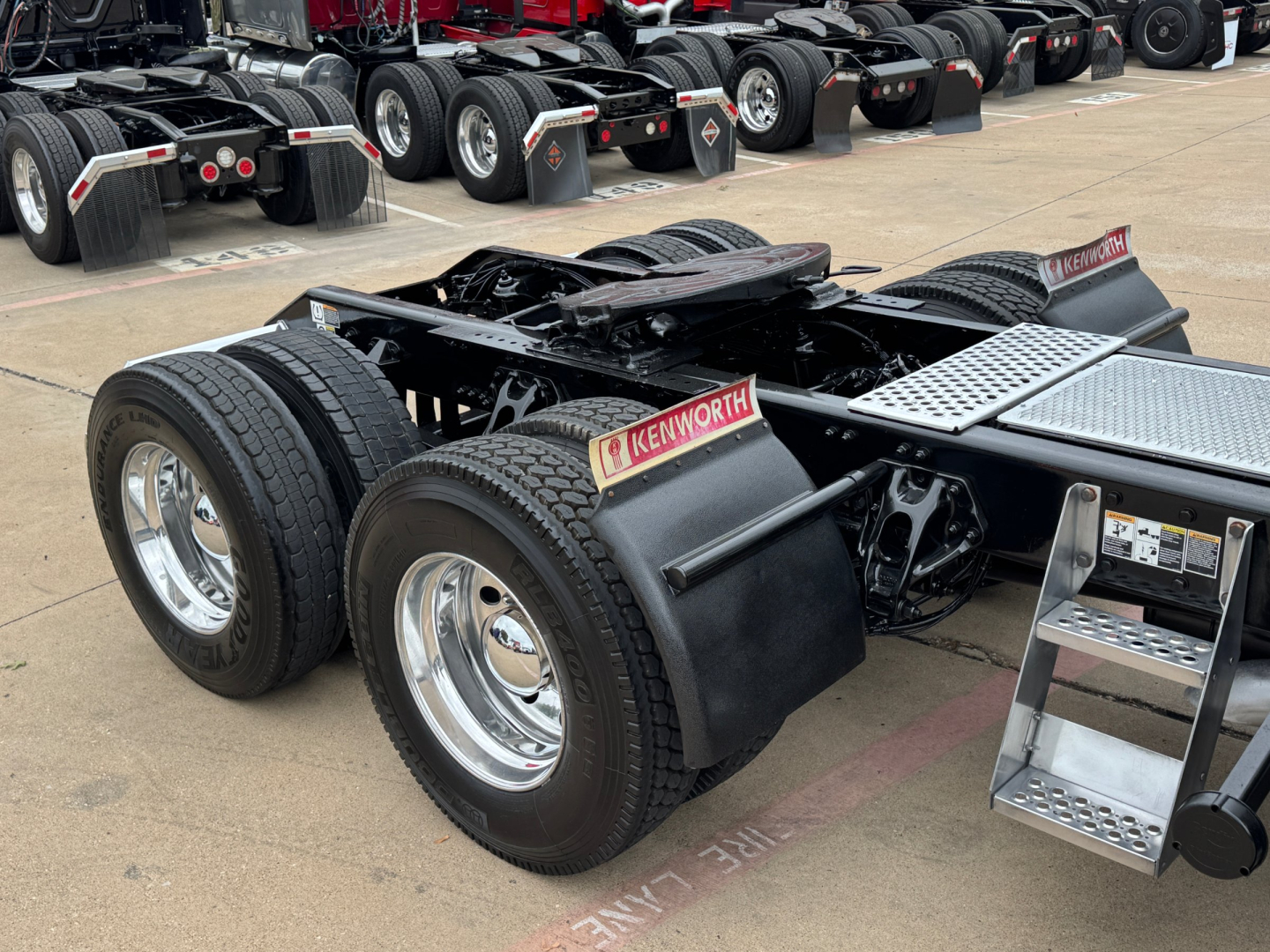 Kenworth T680 rear tractor view showing dual rear axles with Kenworth mudflaps and fuel/step access area. Tires and chrome wheels visible with hydration hoses and chassis components.