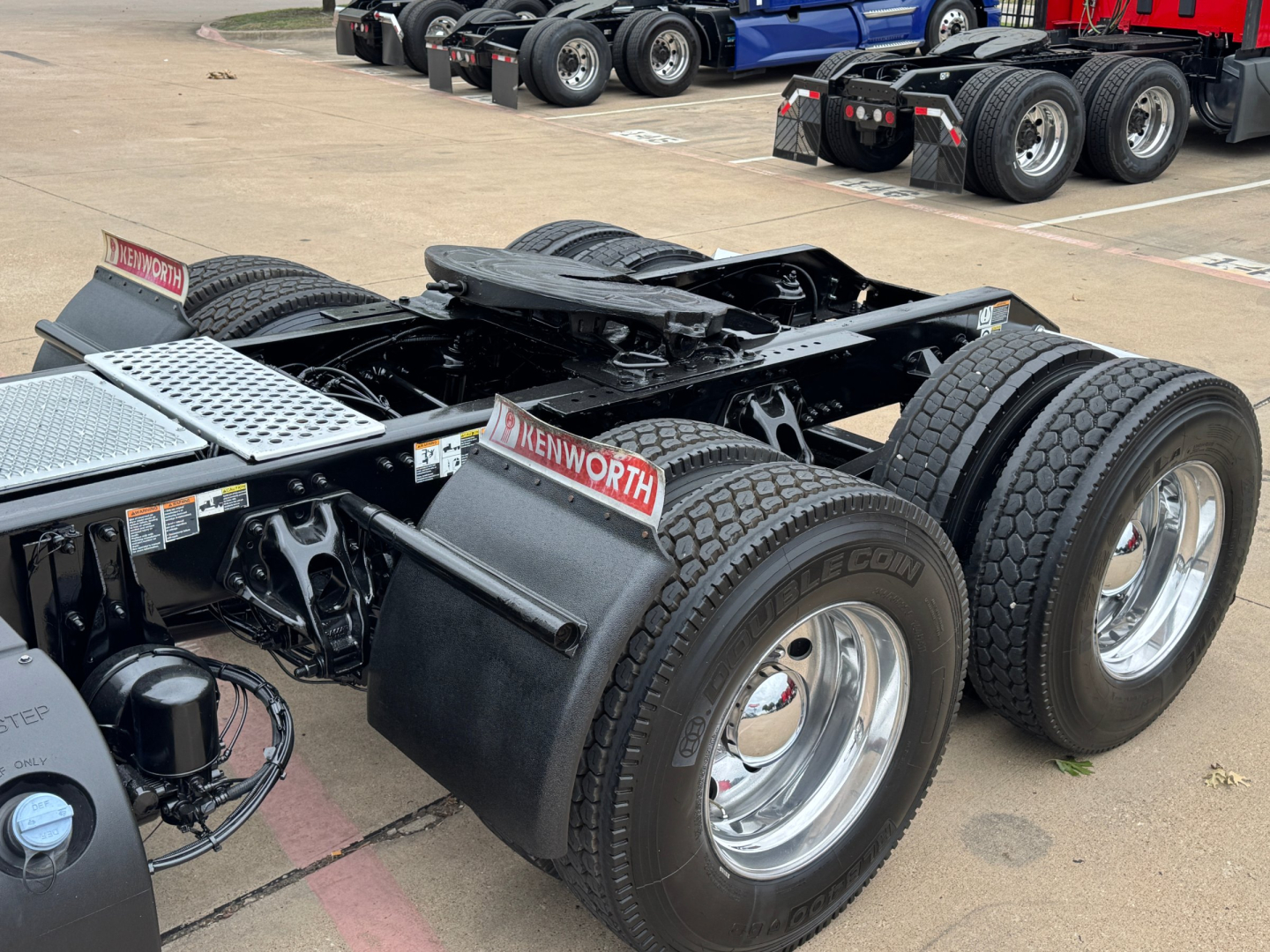 Kenworth truck chassis showing dual rear axles with Kenworth mud flaps and wheels. Visible labels include 'KENWORTH' and various cables and metal steps.