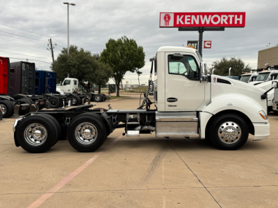 Kenworth T680 tractor side view showing 6x4 chassis, white cab, dual rear axles, fuel tank, and Kenworth dealership sign in background.