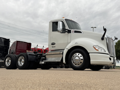 Kenworth T680 conventional daycab truck in off-white with chrome fuel tanks and dual rear axles, side view showing VIN and stock info sticker