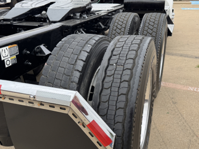 Kenworth truck trailer axles showing tandem rear wheels and tires with chassis rails; warning sticker and metal step visible.