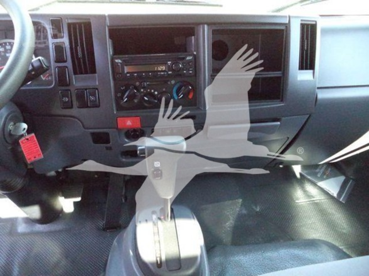 Isuzu truck cab interior showing dashboard with radio, climate controls, gear shifter, and a red hazard button; partial steering wheel and pedals visible.