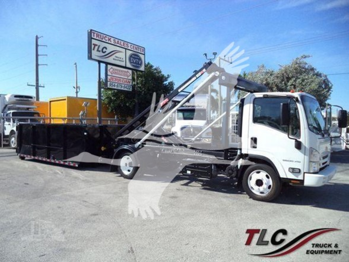 Isuzu NRR roll-off truck with hydraulic lifter and dumping container in yard. Visible text on signs: TLC Truck Sales Center sign in background; on truck cab there may be no legible text. Overall scene shows a white Isuzu NRR roll-off truck 