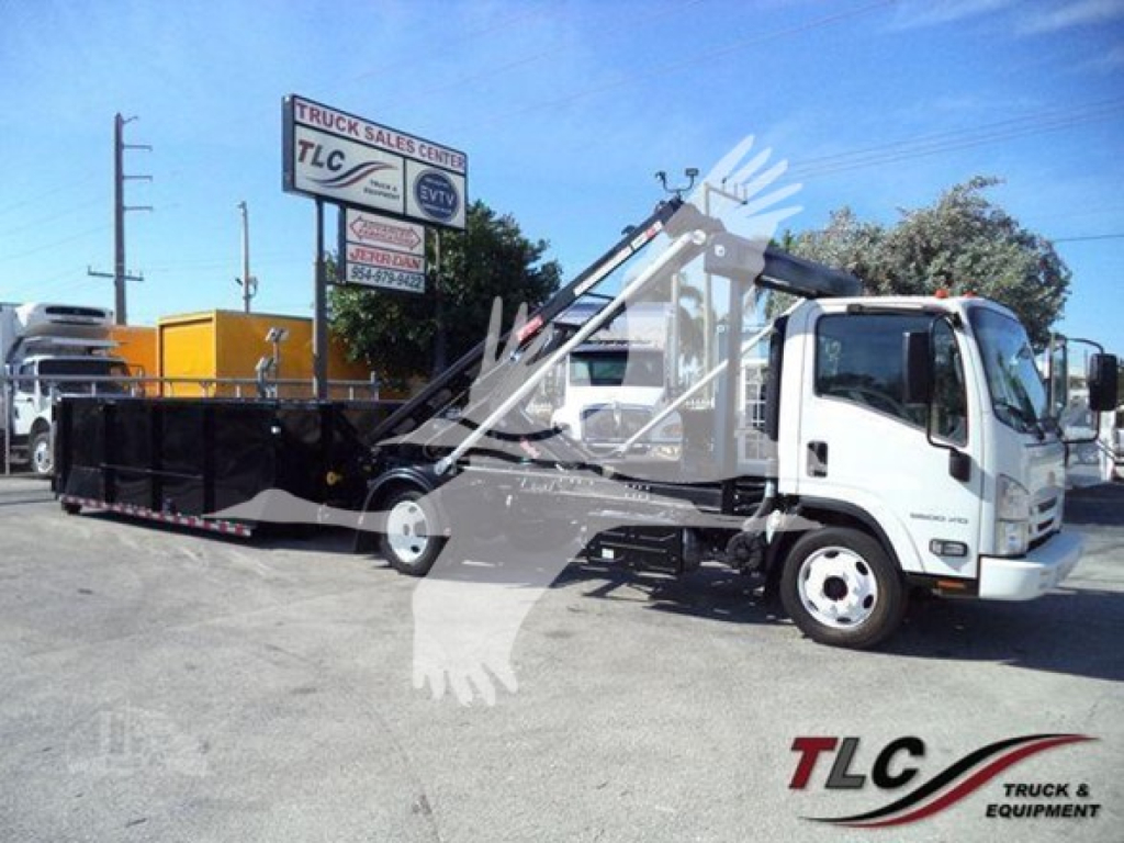 Isuzu NRR roll-off truck with hydraulic lifter and dumping container in yard. Visible text on signs: TLC Truck Sales Center sign in background; on truck cab there may be no legible text. Overall scene shows a white Isuzu NRR roll-off truck 