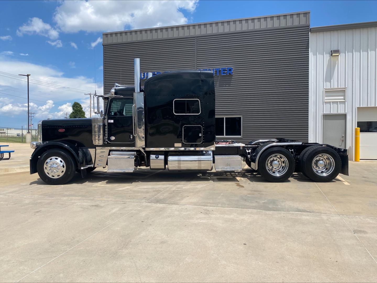 Peterbilt 389 conventional sleeper truck side view, black exterior, dual rear axles, chrome exhaust stacks, and chrome fuel tanks parked in front of a building