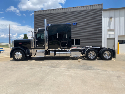 Peterbilt 389 conventional sleeper truck side view, black exterior, dual rear axles, chrome exhaust stacks, and chrome fuel tanks parked in front of a building