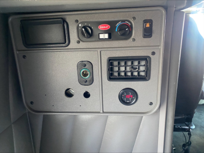 Panel inside Peterbilt truck showing gauges and controls: two round dials, a small rectangular display, a 12V outlet, and an air vent; visible branding on red Peterbilt emblem.
