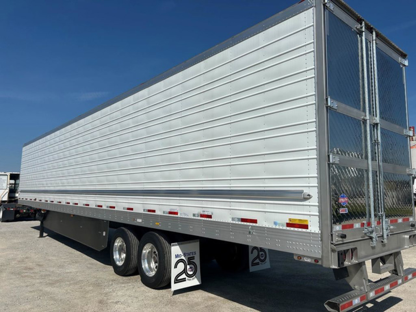 Utility reefer trailer side view showing corrugated aluminum walls and tandem axle configuration, rear doors, reflective strips, and a scale sign under the truck.