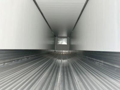 Interior of a refrigerated trailer cargo area with ribbed metal floor and white walls, small rear door opening at far end.