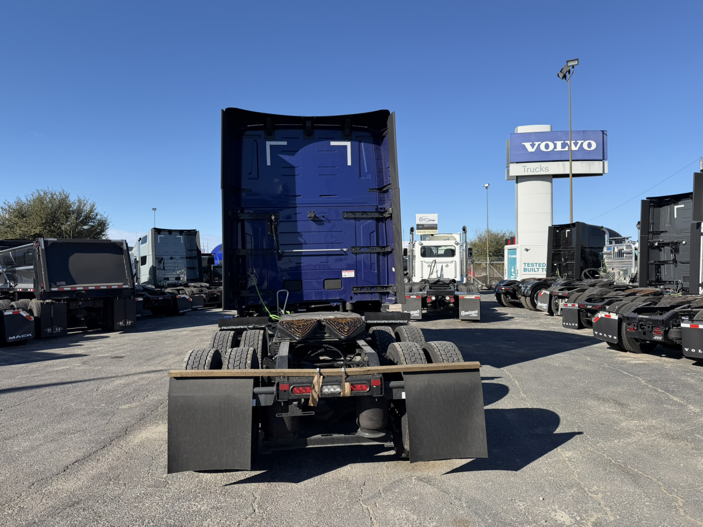 Truck chassis rear view showing fifth-wheel and mounting hardware, blue cab in background, dealership lot with Volvo sign.