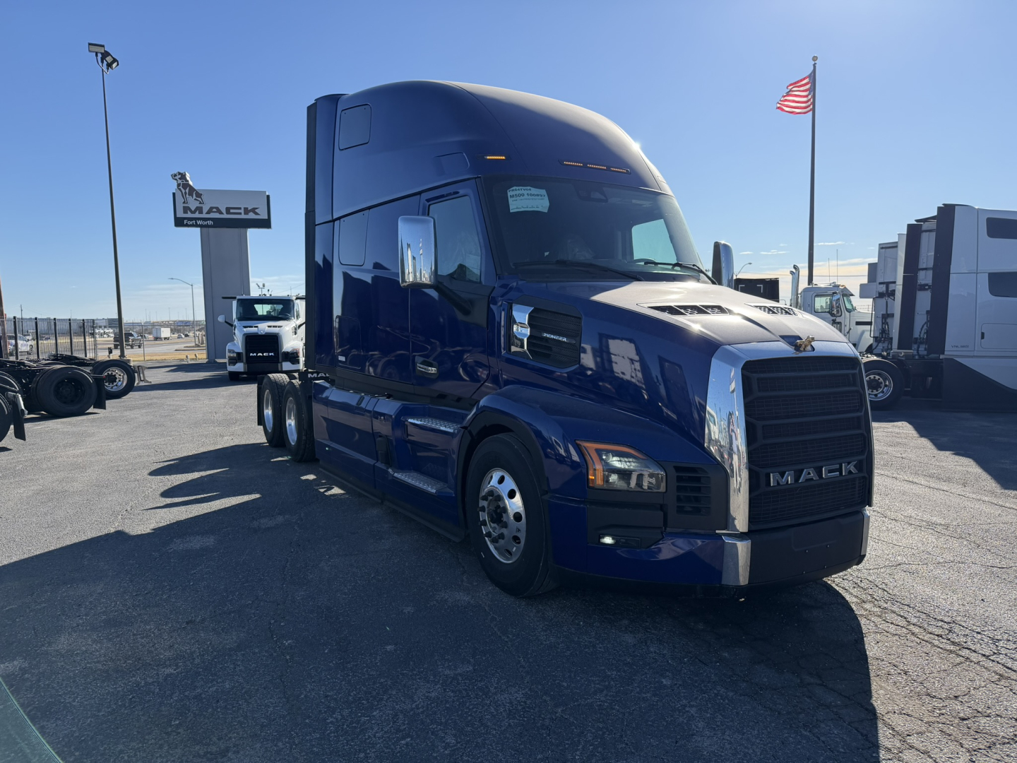 Truck cab side view of a blue 2026 Mack PR64T76H tractor in a dealership lot, front grille with MACK emblem, other trucks in background, American flag.