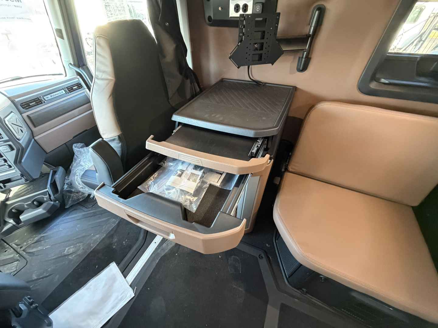 Truck cab interior showing mounted storage shelf and open drawer with paperwork and storage tray, driver seat to left, passenger seat to right, beige and black trim.
