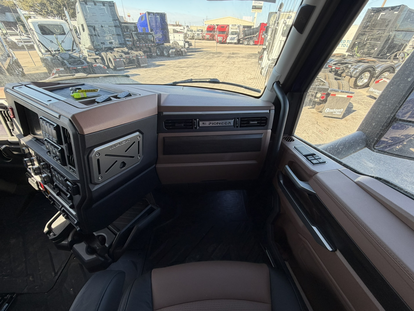 Interior of a new Mack PR64T76H truck cab showing dashboard, steering wheel, and brown upholstery with windows providing external lot view.